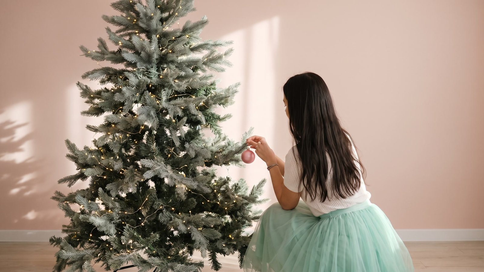 Woman decorating a softly lit Christmas tree in a blush pink room